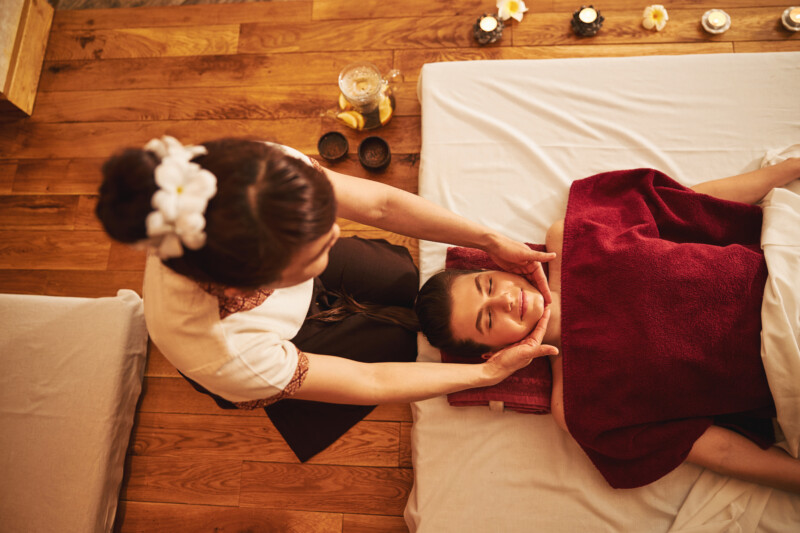 Massage expert standing at the head of mattress with prone female and rubbing her chin with both hands