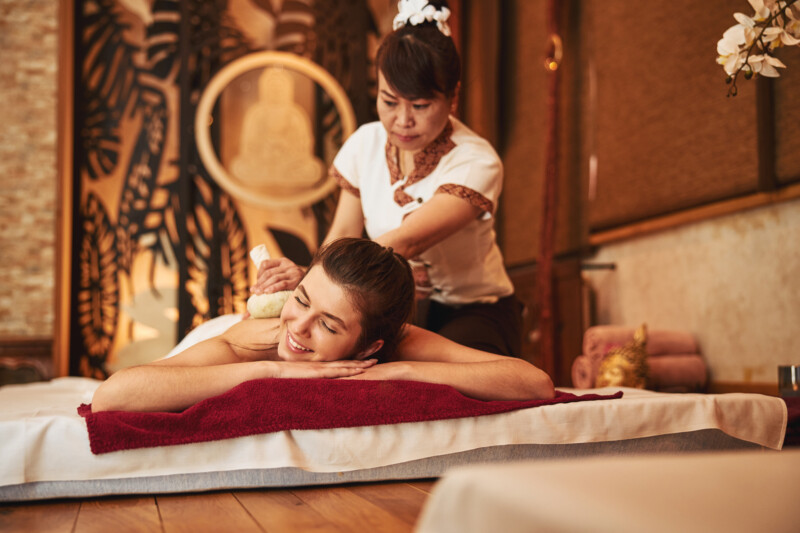 Serious massage parlour Thai professional using herbal compress ball for skin relaxation and tension relief of woman back muscles