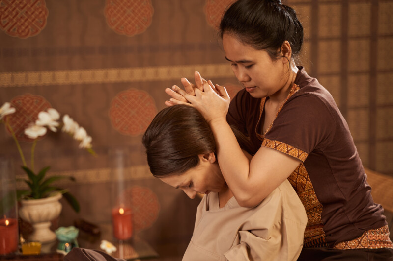 Female massotherapist treating young patient for cervicalgia Experienced mature masseuse stretching neck muscles of her client during Thai massage session