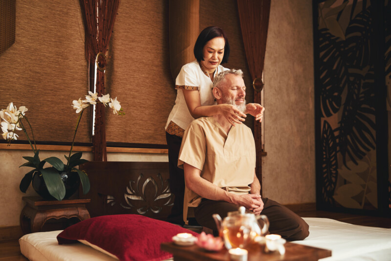 Grizzled bearded male is relaxing in oriental studio while woman is healing his neck with hands